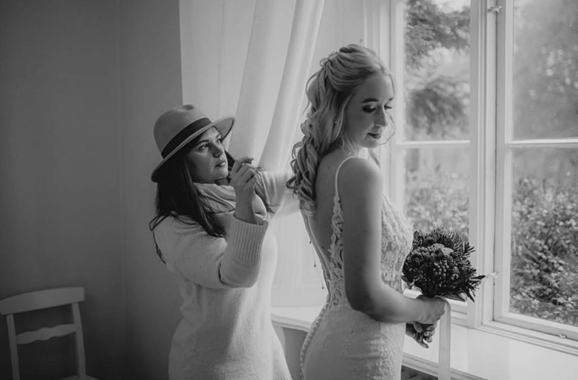 Eine Frau in einem Hochzeitskleid hält einen Blumenstrauß und steht am Fenster, während eine andere Frau ihrem Haar den letzten Schliff verleiht.
