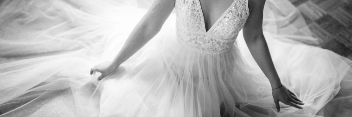Eine Frau in einem Hochzeitskleid hält einen Blumenstrauß und steht am Fenster, während eine andere Frau ihrem Haar den letzten Schliff verleiht.